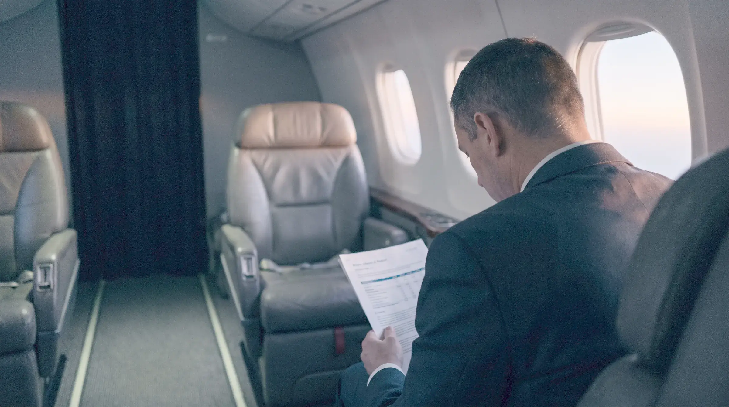 Ejecutivo sentado junto a la ventanilla de un avión, visto de espaldas, leyendo un documento al amanecer